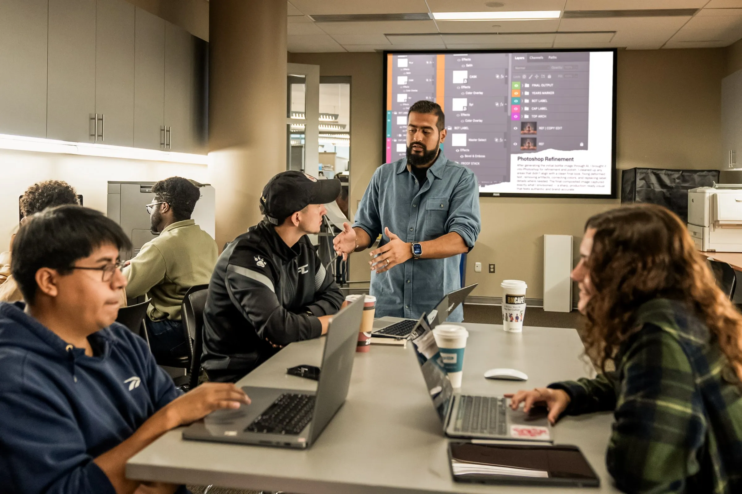 Sahil Patel directing a critique with students at laptops, a Photoshop layer stack projected on the screen behind him